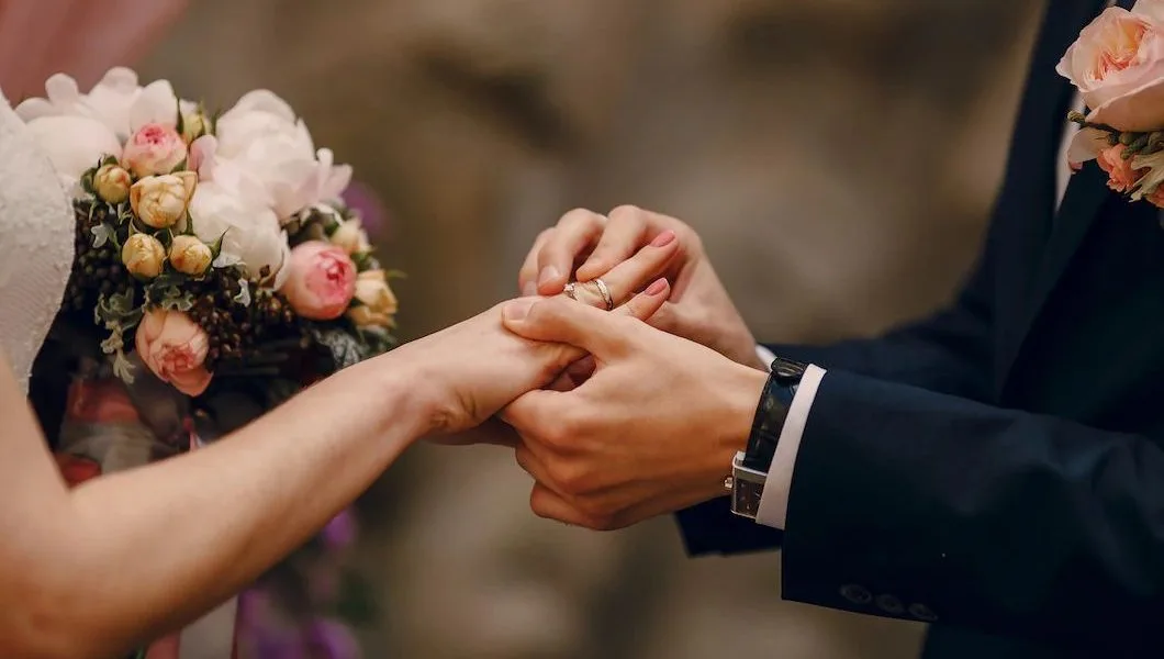 Noivos de mãos dadas trocando alianças na cerimônia de casamento, com buquê de rosas em tons de rosa e pêssego